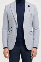 Uomo che indossa un blazer grigio chiaro, un dolcevita blu navy e pantaloni scuri su uno sfondo bianco