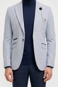 Uomo che indossa un blazer grigio chiaro, un dolcevita blu navy e pantaloni scuri su uno sfondo bianco